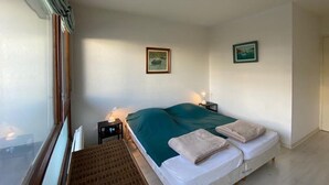 Ein Zimmer mit einem Einzelbett, einem Fenster, einem Nachttisch mit Lampe und zwei gerahmten Bildern an der Wand.
