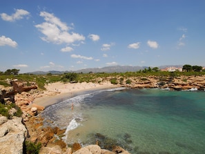 A coastal beach with clear turquoise waters, rocky shoreline, and distant mountains.