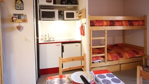 Una habitación compacta con literas, una kitchenette con microondas y cafetera, una pequeña mesa con un plato y un cuchillo, y una silla.