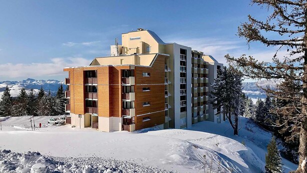 Un hotel moderno con fachada de madera, varios balcones y vistas a montañas nevadas.