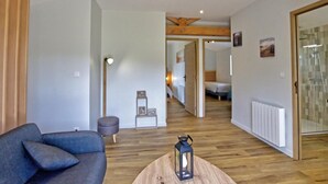Un soggiorno moderno con un divano, un tavolino rotondo con una lanterna e un pavimento in legno. Si apre un corridoio che conduce a una camera da letto con un letto e un bagno con doccia.