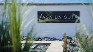 Un letrero que dice "ASA DA SUTA" en una pared con un paisaje rocoso y algo de vegetación en primer plano.