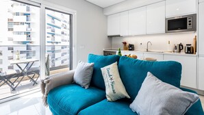 Un appartement moderne avec un canapé de couleur turquoise, une kitchenette et un balcon offrant une vue sur un immeuble d’habitation.