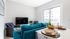 Un salon moderne avec un canapé de couleur turquoise, un téléviseur à écran plat, une table à manger dressée avec des bols et une bouteille, et une vue sur le paysage urbain à travers de grandes fenêtres.