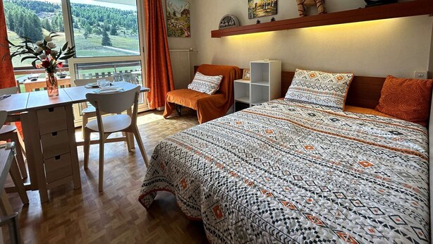 A bedroom with a large bed, a desk with a chair, a small table with a vase of flowers, and a window with a view of trees.