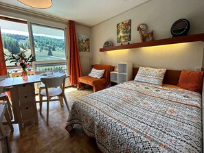 Une chambre avec un grand lit, un bureau avec une chaise, une petite table avec un vase de fleurs et une fenêtre donnant sur des arbres.