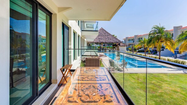 Un balcon doté d’une balustrade en verre donnant sur une piscine, un parasol en chaume et une vue sur un bâtiment d’un complexe hôtelier.