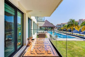Un balcon doté d’une balustrade en verre donnant sur une piscine, un parasol en chaume et une vue sur un bâtiment d’un complexe hôtelier.