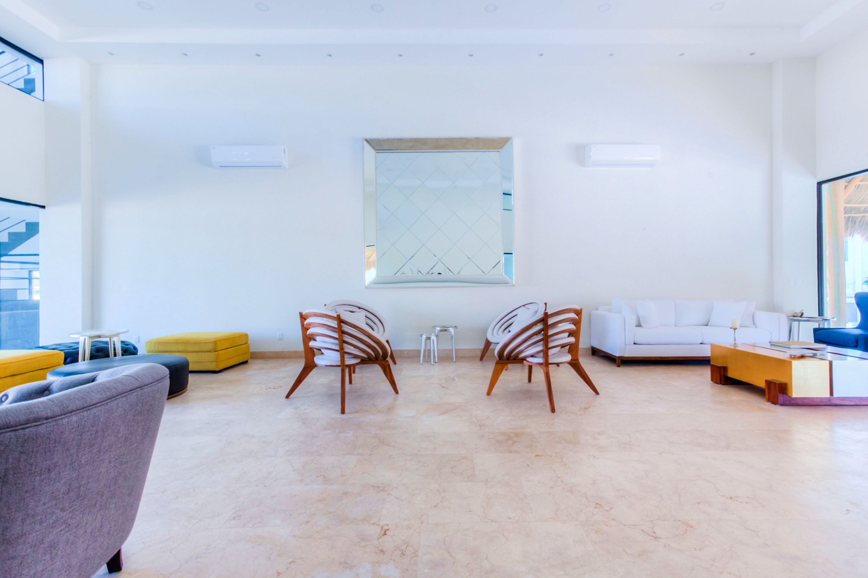 A modern living room with a white sofa, a grey armchair, and two wooden chairs. There is a yellow ottoman, a small white table, and a blue chair. A large window with a geometric pattern is visible on the wall.