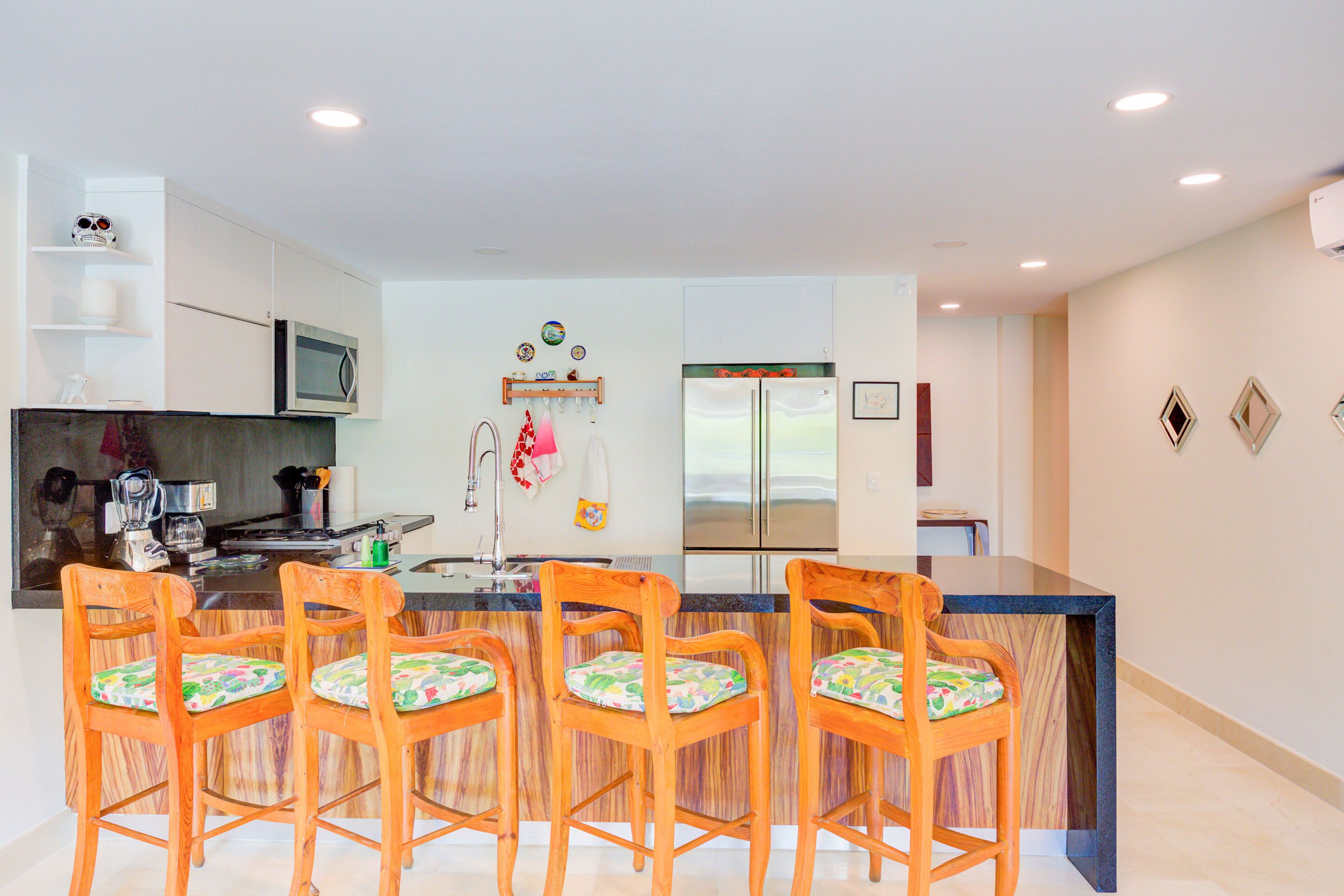 A modern kitchen with a central island, wooden chairs with colorful cushions, a built-in microwave, and a refrigerator.