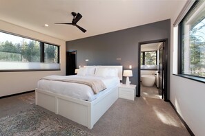 A modern bedroom with a large bed, bedside lamps, a ceiling fan, and a view of the outdoors.