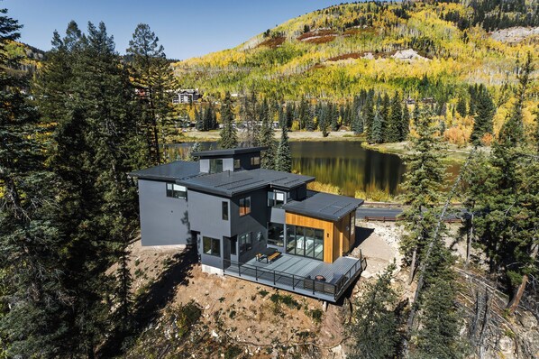 A modern two-story house with a deck overlooking a lake, surrounded by dense forest and autumn foliage.