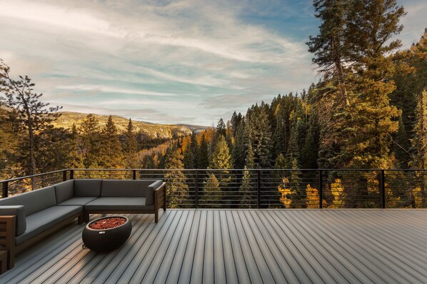 A deck with a sofa and a fire pit, overlooking a forested area. - New Lake Front Home Across from Purgatory - Multiple Decks, Views & Ping Pong (Durango)
