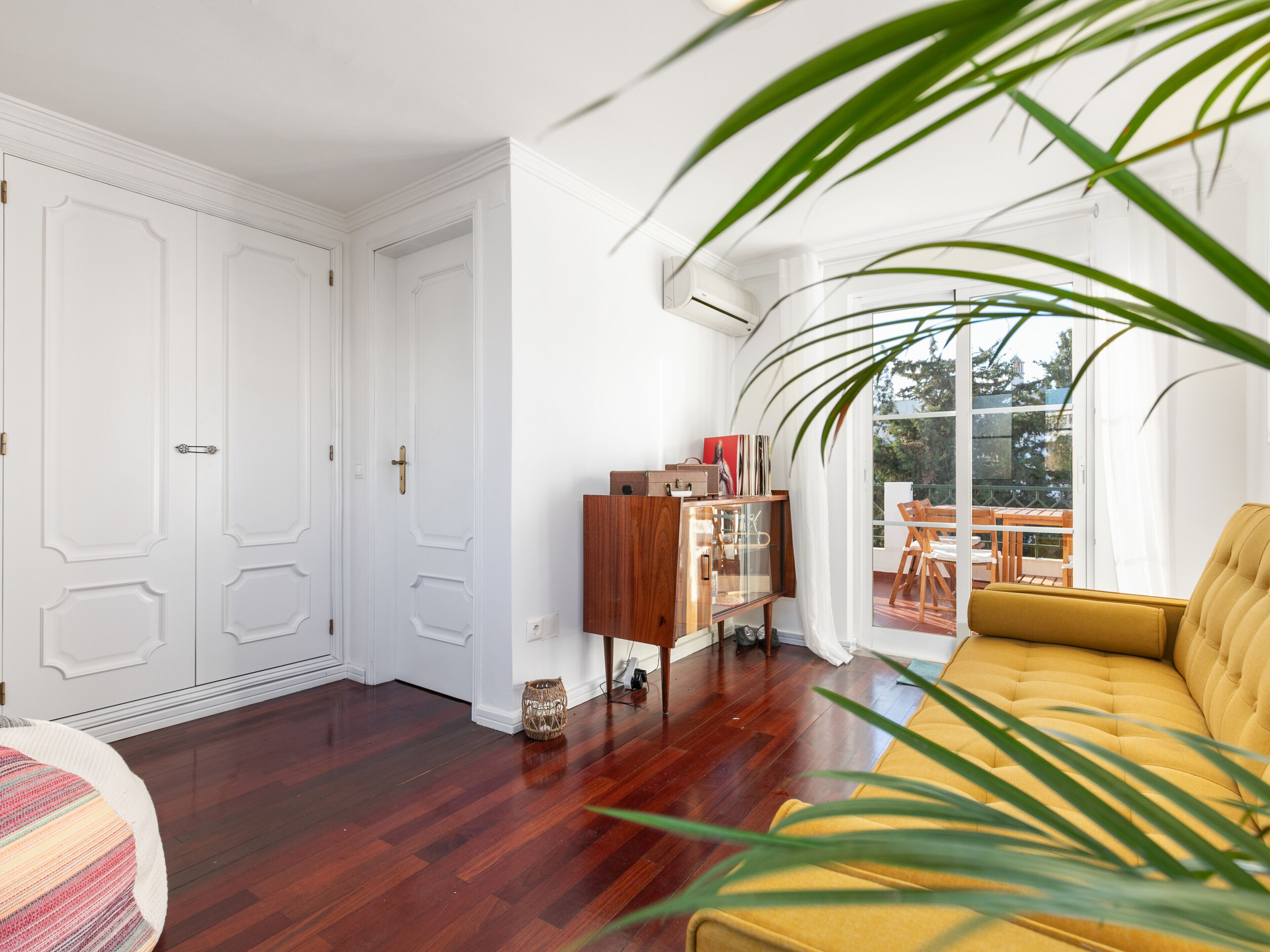Una habitación con piso de madera, un sofá amarillo, un armario de madera y un ventanal con vistas al exterior.