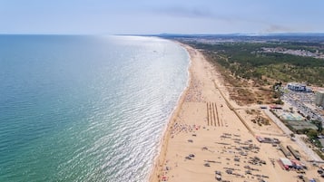 Vista aérea de una playa con una extensa franja de arena, algunas embarcaciones y un pequeño edificio a la derecha.