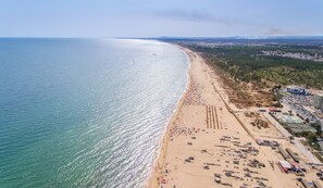 An aerial view of a beach with a long stretch of sand, a few boats, and a distant cityscape.