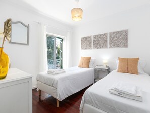 A hotel room with two beds, a window with a view, a bedside table with a lamp, and decorative wall art.