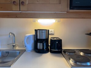 A kitchen with a stainless steel sink, a white electric kettle, a black coffee maker, a black toaster, and a stove with four burners.