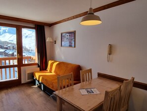 A cozy room with a wooden table and chairs, a sofa, a window with a view of snow-covered mountains, and a mounted telephone.