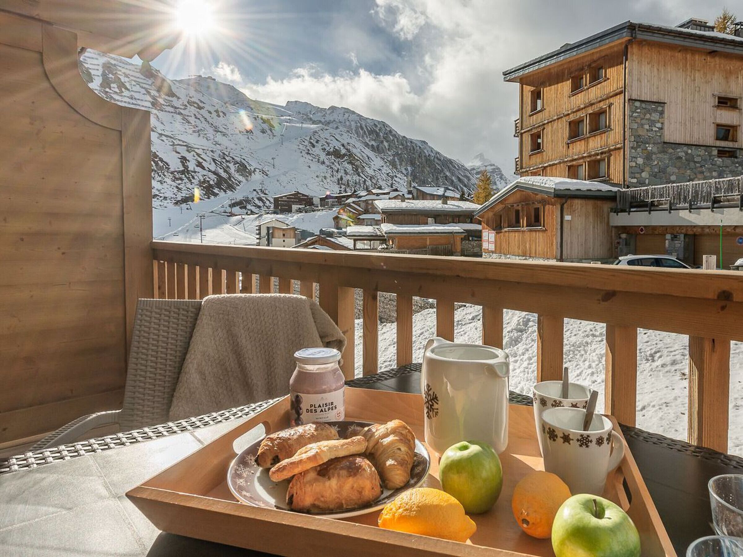 A wooden deck with a table set for breakfast, overlooking a snowy mountain village.