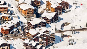 A snow-covered mountain resort with multiple wooden buildings, ski lifts, and a clear sky.