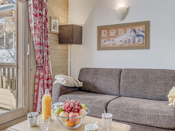Ein gemütliches Wohnzimmer mit einem grauen Sofa, einem Glastisch mit Obst und Getränken, einem roten blumigen Vorhang und einem gerahmten Kunstwerk an der Wand.