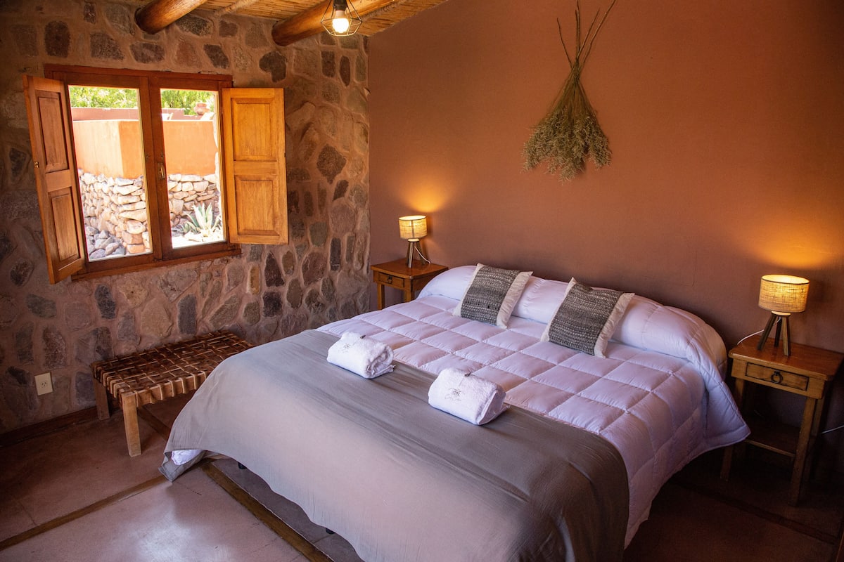 A bedroom with a stone wall, a wooden bench, a bed with white bedding, and two bedside lamps.