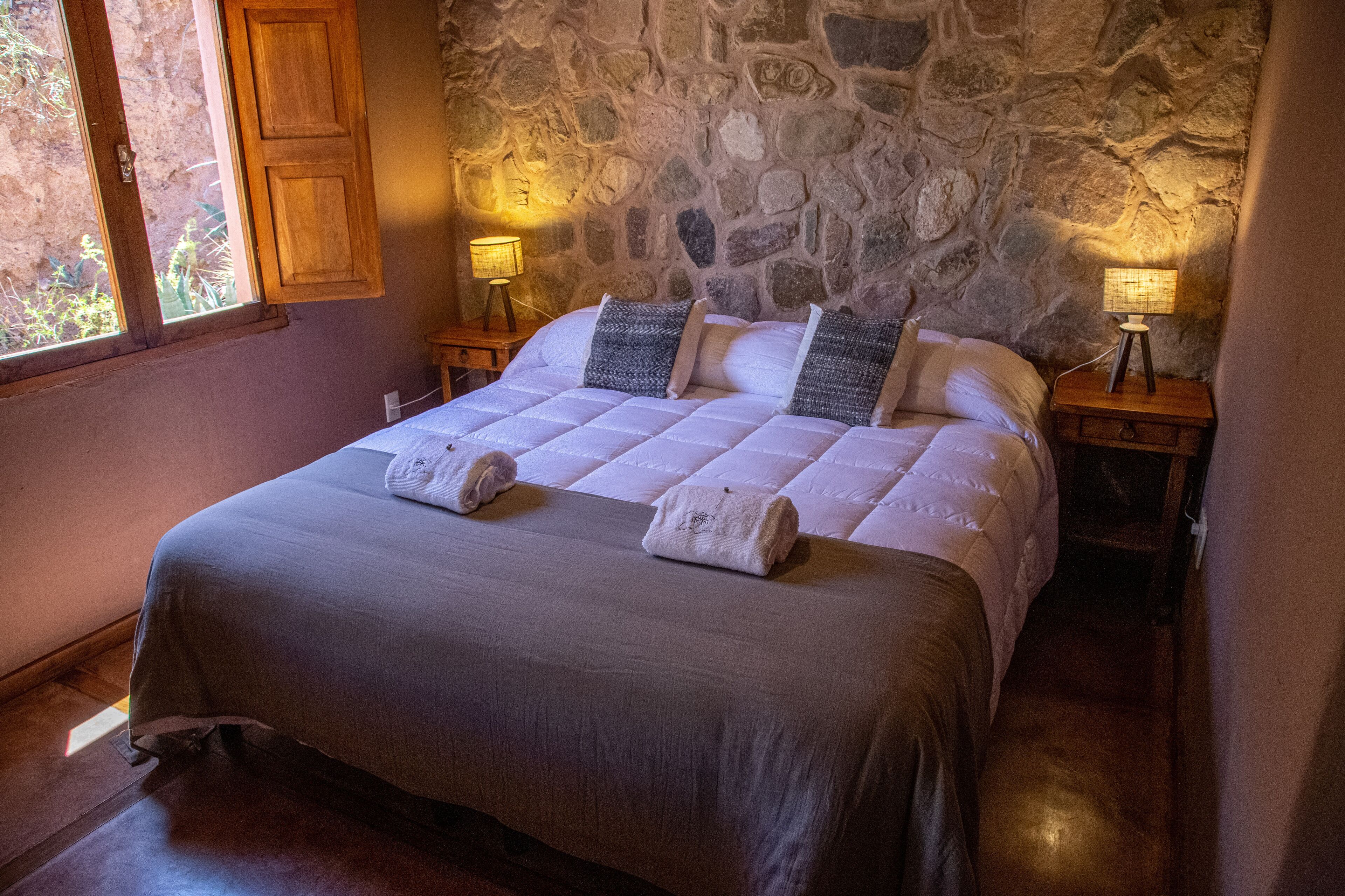 Un letto con una parete in pietra alle spalle, due comodini con lampade e una finestra con persiana in legno.
