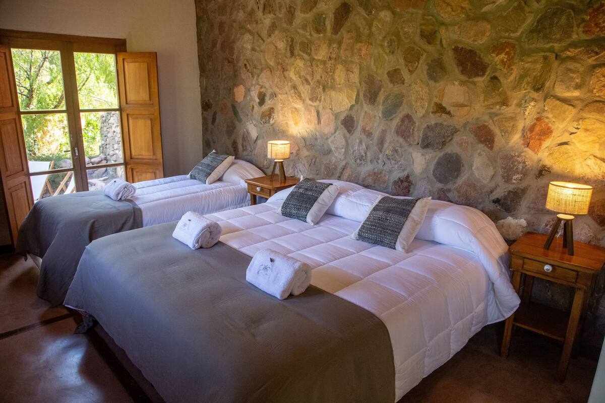 A room with two beds, stone walls, and a window with a view of greenery.