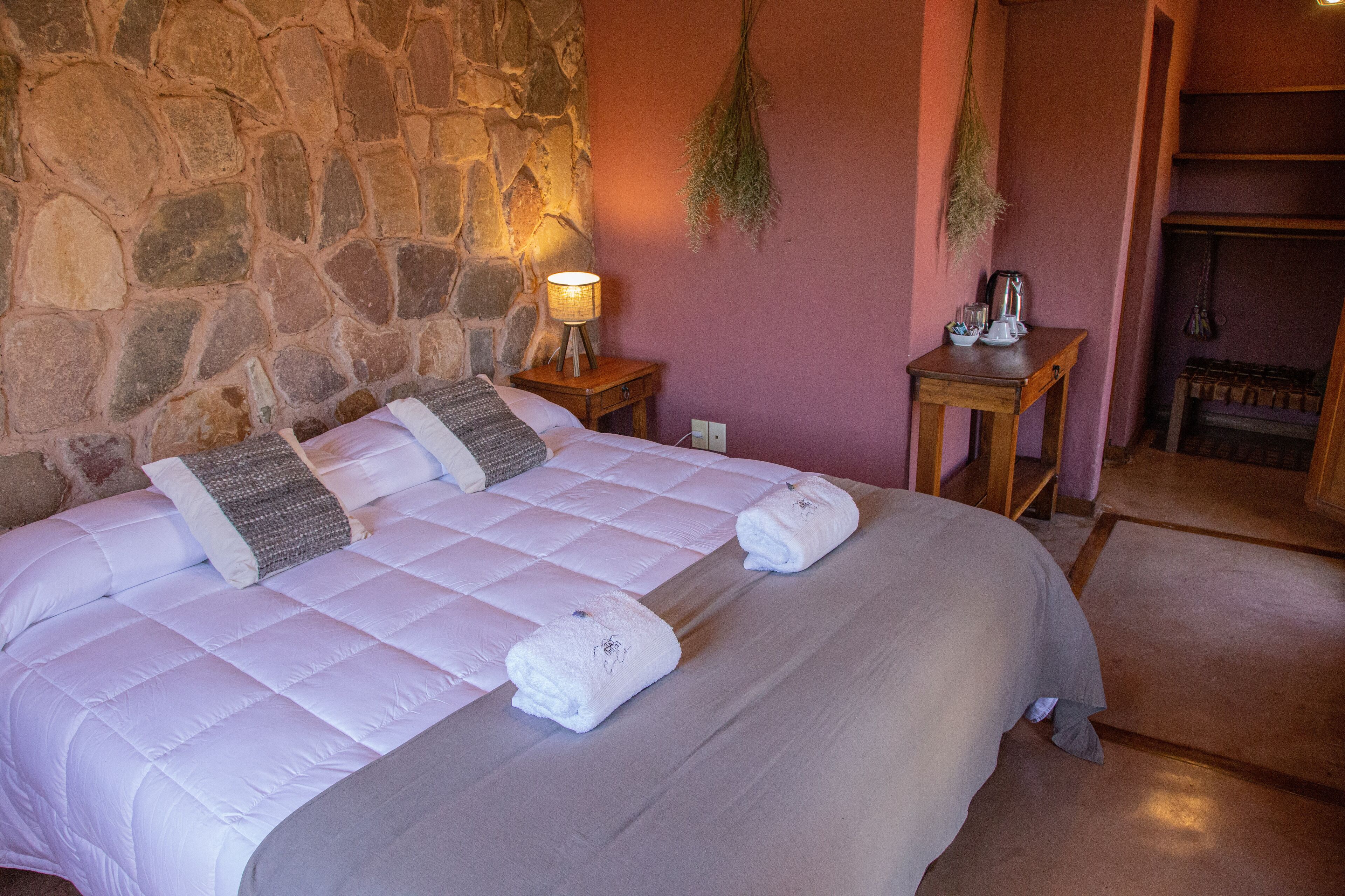 Una cama con una pared de piedra de fondo, una mesita de noche con una lámpara y una pequeña mesa con un hervidor.