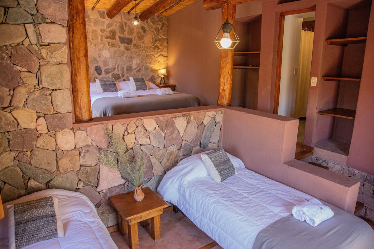 A bedroom with stone walls, a wooden ceiling, and a large mirror reflecting the bed. There are two beds with white linens and a small wooden nightstand with a vase.