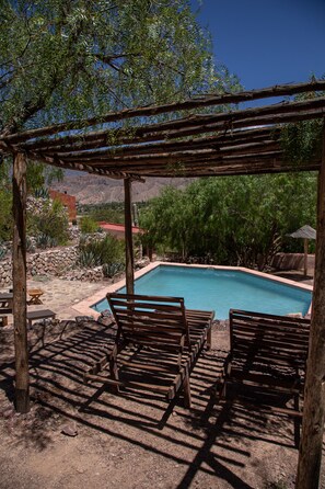 A pool area with wooden chairs and a pergola, surrounded by greenery and a mountainous backdrop. - CasaCalma Hotel Boutique (Tilcara)