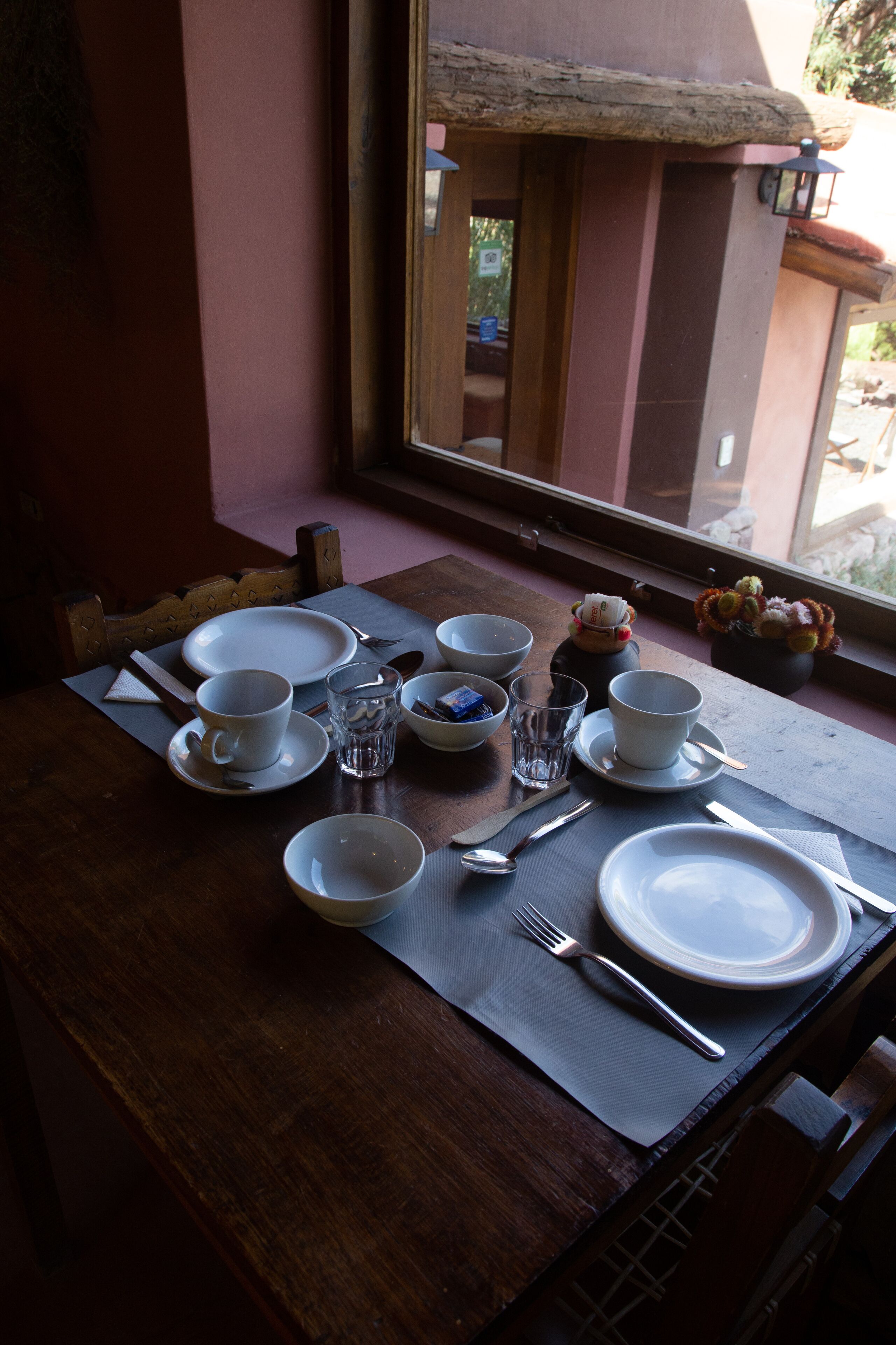 A dining table set with white plates, bowls, and cutlery, positioned near a window with a view of the outdoors.