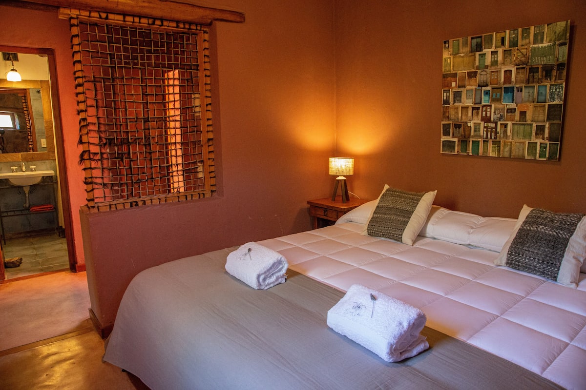 A hotel room with a bed, a nightstand with a lamp, a wall art piece, and a window with a wooden blind.