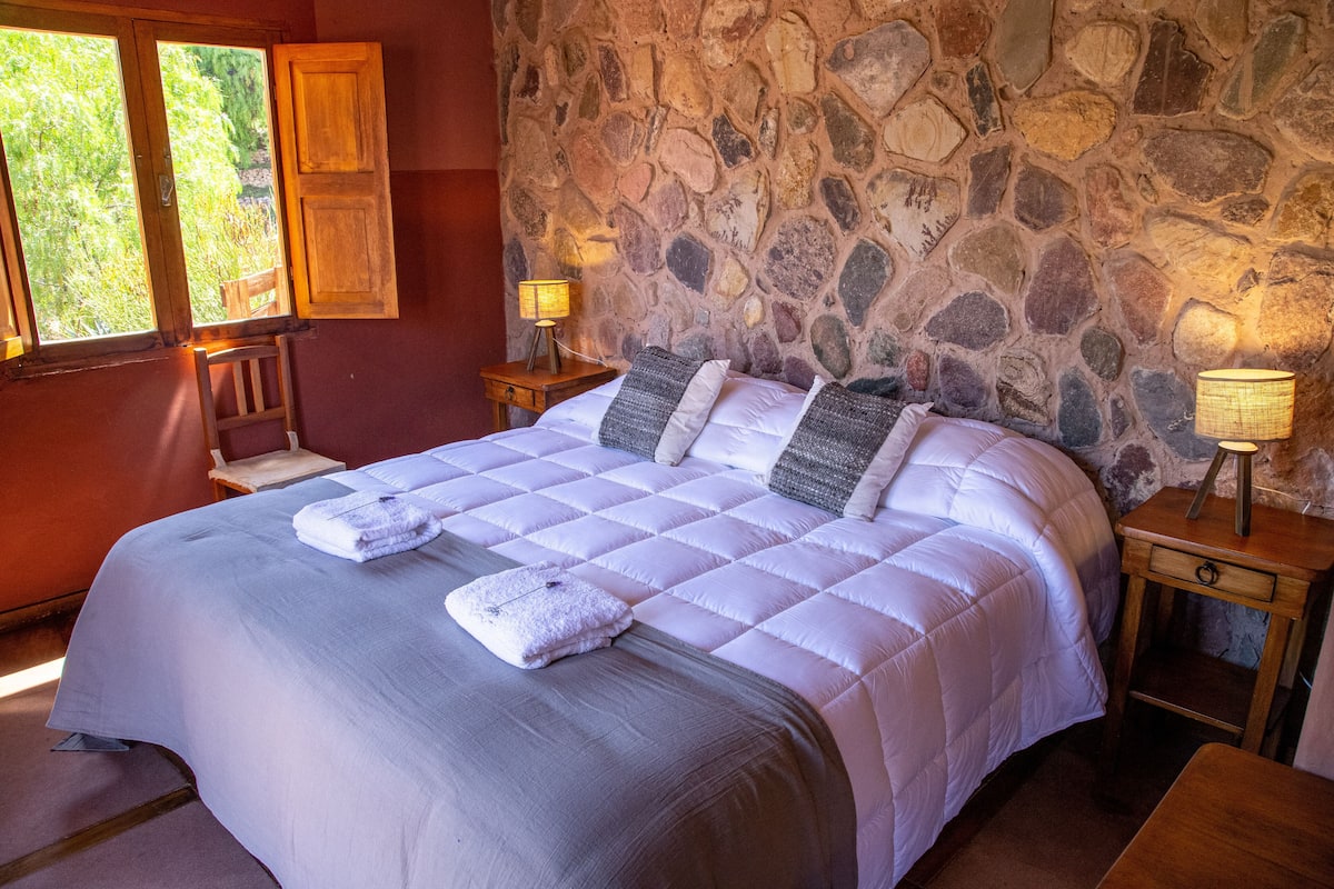 A neatly made bed with a stone wall backdrop, two bedside tables with lamps, and a window with wooden shutters.