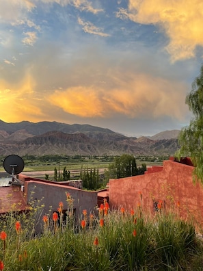 A mountainous landscape with a sunset, featuring a building with a satellite dish and red walls, surrounded by green vegetation and orange flowers. - CasaCalma Hotel Boutique (Tilcara)