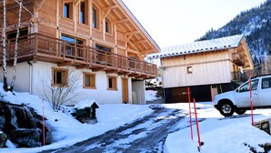 A wooden chalet with a balcony, a garage, and a snow-covered driveway.