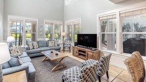 Ein geräumiges Wohnzimmer mit grauem Sofa, gemusterten Sesseln, einem Couchtisch und einem Fernseher auf einem Holzständer. Große Fenster bieten Blick ins Freie.