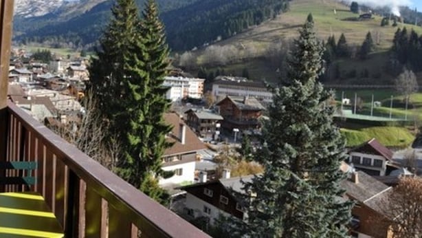 Un pueblo de montaña con picos nevados al fondo, una combinación de edificios residenciales y comerciales, y un imponente pino en primer plano.