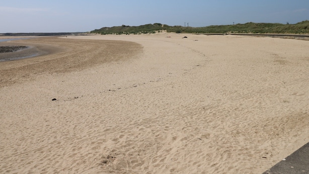 Een breed zandstrand met zachte golven, een heldere lucht en duinen in de verte.