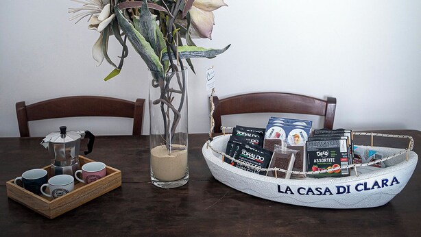 Een eettafel met een koffiezetapparaat, een dienblad met koffiekopjes, een vaas met bloemen en een mandje met de tekst 'LA CASA DI CLARA' dat koffieproducten bevat.