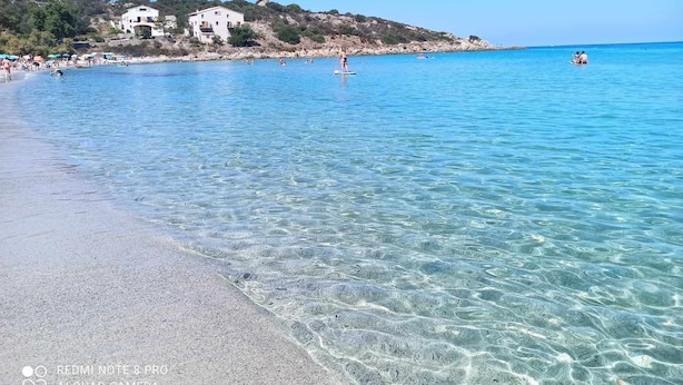 Een strand met helder water, enkele mensen en gebouwen op de achtergrond.