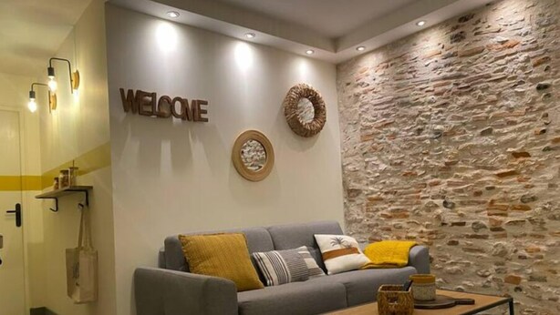 Een moderne woonkamer met een grijze bank, een bijzettafeltje, een stenen muur en een bord met de tekst 'WELCOME'.