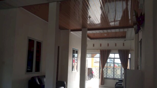 A spacious room with a high ceiling, a large window with curtains, a black leather sofa, and a tiled floor. - Hotel O Ananda Homestay Syariah (Pekanbaru)