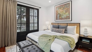 A bedroom with a large bed, a window with curtains, a ceiling fan, a wall-mounted artwork, bedside lamps, and a bedside table with a stack of books. - 5BR Victorian Carriage House | Near Forsyth Park (Savannah)