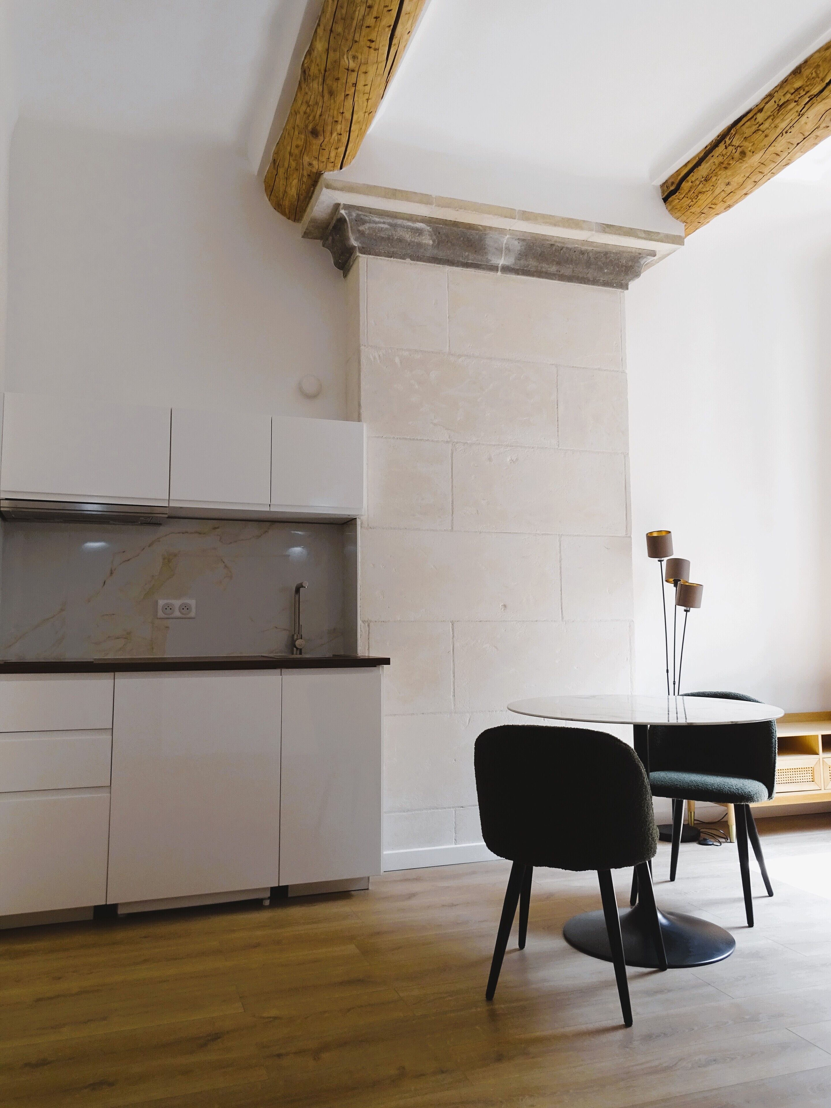 Una cocina moderna con armarios blancos, una encimera de mármol y techo de madera. Hay una mesa redonda con dos sillas y una lámpara de pie alta.
