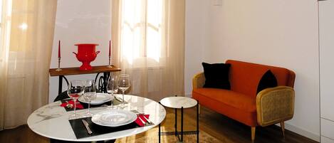 Un comedor con una mesa redonda preparada para cuatro personas, un florero rojo y un pequeño sofá naranja.
