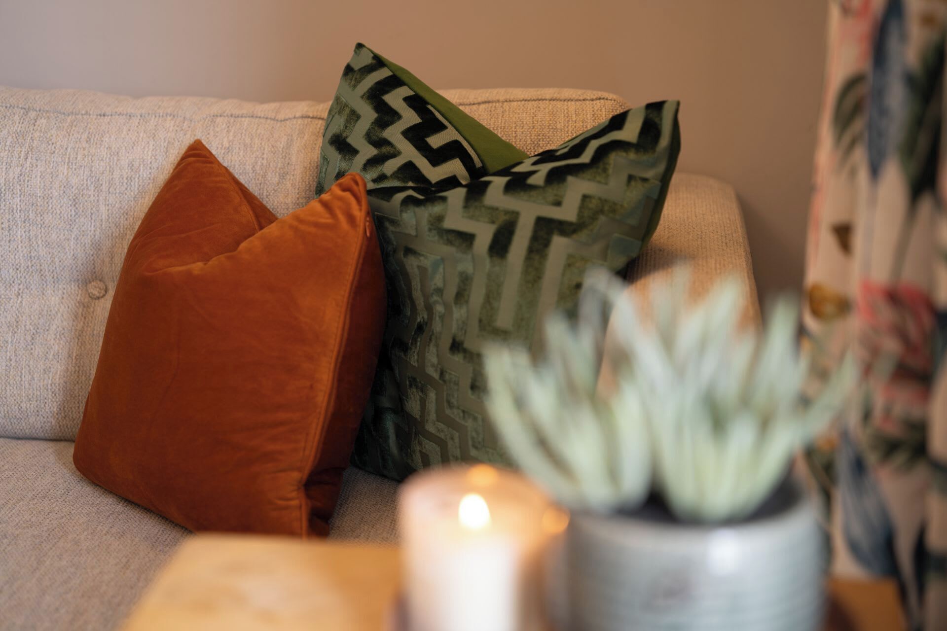 Ein beiges Sofa mit zwei Kissen, eines orange und eines mit einem geometrischen Muster, und eine Topfpflanze im Vordergrund.