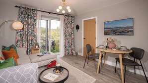 Ein gemütliches Wohnzimmer mit einer Couch, einem Couchtisch und einem Essbereich mit Tisch und Stühlen. Ein Fenster mit Vorhängen, eine Wanduhr und ein Gemälde sind ebenfalls zu sehen.