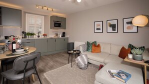 Ein modernes Wohnzimmer mit einer Couch, einem Couchtisch und einem Essbereich mit Stühlen. Es gibt eine Küche mit einer Mikrowelle und einem Spülbecken.